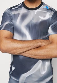 Camisa atlética de manga corta en tonos de gris con patrones abstractos en blanco, tela texturizada, cuello negro y pequeño detalle del logo en azul.