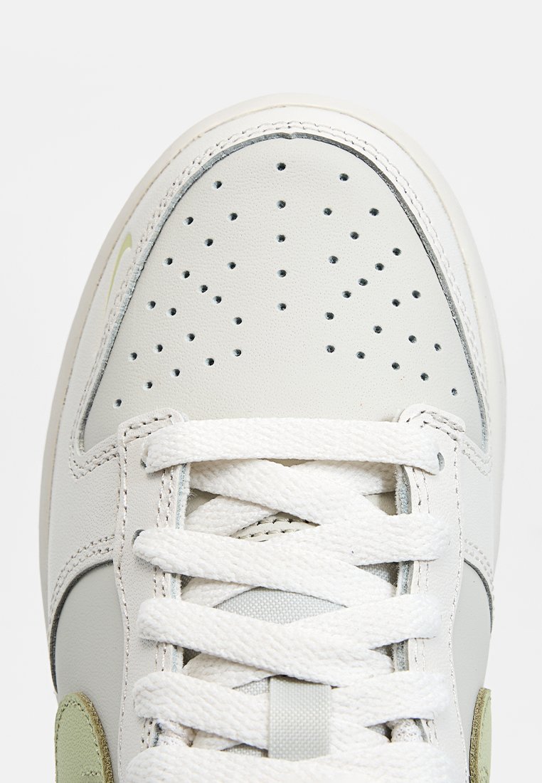 Sneaker en cuir blanc avec une boîte de orteil perforée, des lacets ronds blancs et des accents verts sur le côté. Texture lisse avec des éléments de design courbés.