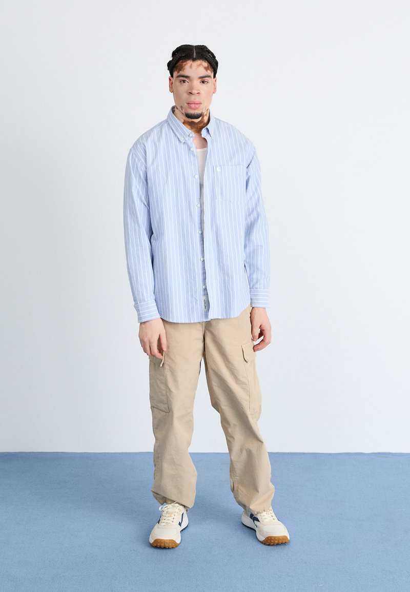 Hellblau-gestreiftes Button-Up-Hemd, beigefarbene Cargo-Hosen und beigefarbene Sneaker mit dunklen Akzenten. Schlichter Hintergrund mit einem blauen Boden.