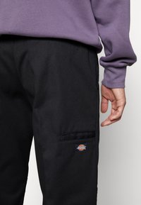 Svarta cargo-byxor med en slät yta, med en sidoficka och en liten flerfärgad logotyp-patch på vänster ben.