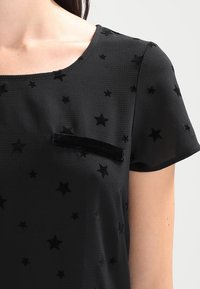 Top negro de manga corta con un patrón de estrellas y un bolsillo de terciopelo en el pecho. La tela tiene un acabado texturizado con un suave caído.
