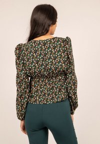 Blouse noire à motifs floraux avec des manches bouffantes, ornée de petites fleurs multicolores, taille froncée et décolleté rond. Portée avec un pantalon vert.