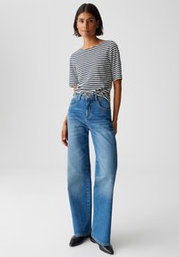 Femme debout portant une chemise à rayures noires et blanches à manches courtes rentrée dans un jean bleu taille haute à jambes larges et des chaussures noires à talons pointus.