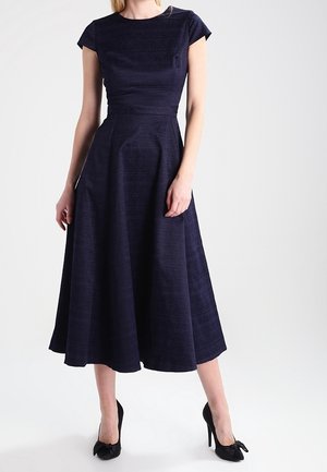 Frau trägt ein marineblaues, kurzärmeliges, knielanges A-Linien-Kleid mit schwarzen High Heels, die Schleifenakzente aufweisen.