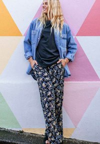 Veste en denim oversize, t-shirt noir et pantalon ample à motifs floraux en bleu marine avec des fleurs jaunes et blanches, devant un mur géométrique coloré.