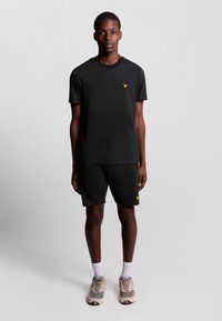 T-shirt nera e pantaloni corti coordinati, entrambi in cotone. Un logo giallo sul petto e sui pantaloni. Il modello indossa scarpe da ginnastica grigio chiaro e calze bianche.