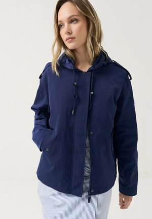 Ung kvinde med blondt hår iført en navy hættetrøje over et lyseblåt og hvidt striped skørt, der står mod en ensfarvet baggrund.