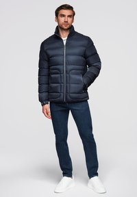 Marineblå pufferjakke med quiltet design, høj krave og lynlås foran. Kombineret med mørke jeans og hvide sneakers. Glat tekstur.