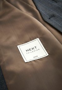 Grau zugeschnittene Anzugjacke mit glattem braunem Futter und einem weißen Etikett mit der Aufschrift "NEXT TAILORING 40R." Sichtbare Nähte sind vorhanden.