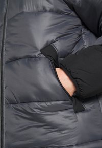 Pufferjakke i mørkegrå med quiltet design. Har en sort stoflomme og blød, isoleret tekstur. Lynlås synlig på lommesiden.