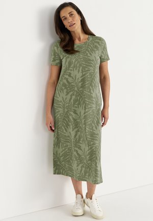 Kvinde stående mod en hvid væg iført en grøn midi kjole med blad mønster og hvide sneakers, kiggende let fremadbøjet.