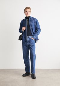 Camisa de denim y vaqueros a juego en azul medio, sobre una camiseta de cuello alto negra. Bolso cruzado negro y botas gruesas negras. Diseño sencillo.