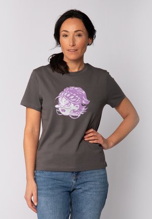 Vrouw met donker haar in een paardenstaart die een grijs T-shirt met een paarse cartoongezichten erop en een blauwe spijkerbroek draagt, staand met één hand op haar heup.
