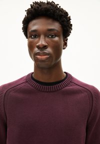 Lila gestrickter Pullover mit einem gerippten Halsausschnitt, detaillierten Schulternähten und einer glatten Textur. Figurbetontes Design, getragen über einem schwarzen Shirt.