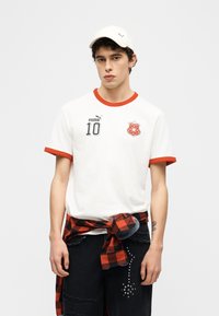 Νεαρός άνδρας με λευκό αθλητικό πουκάμισο Puma με κόκκινη επένδυση και αριθμό 10, κόκκινο πουκάμισο καρό δεμένο στη μέση, μαύρο παντελόνι με τρουκς και λευκό καπέλο.