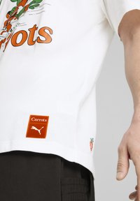 Balta kokvilnas T-kreklu ar oranžu "Burkāni" grafiku, izšūtu burkānu detaļu apakšmalā un sarkanu logo uzšuvumu ar Puma zīmolu.