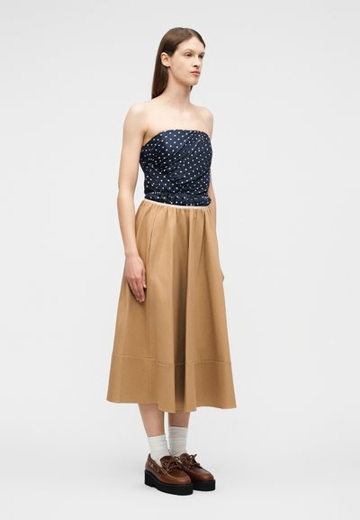 Γυναίκα που φοράει μπλε χωρίς τιράντες τοπ με πουά, καφέ midi φούστα, λευκές κάλτσες και καφέ πλατφόρμες, στέκεται μπροστά σε απλό φόντο.