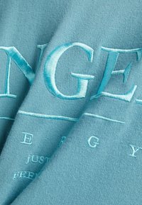 Turkos tyg med upphöjd, ljusblå broderi av text. Slät struktur, vikt med framträdande designdetaljer på en mjuk bakgrund.