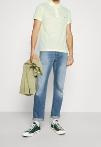 Světle žluté polo tričko, modré džíny, zelené tenisky a olivový svetr složený v ruce. Neformální outfit s ohrnutými rukávy a detailem loga.