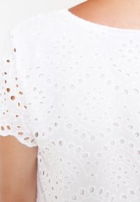 Haut en coton blanc présentant un travail de découpe complexe et des motifs brodés. Encolure ronde et manches courtes à bords festonnés.