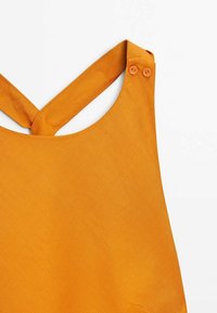 Vêtement sans manches jaune moutarde avec de larges bretelles croisées dans le dos, maintenu par deux boutons assortis sur une épaule.