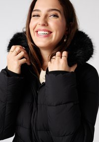 Zwarte pufferjack met een nepbont capuchon, voorzien van een ritssluiting, gequilt textuur en contrasterende binnenmanchetten, gedragen door een persoon met ringen.