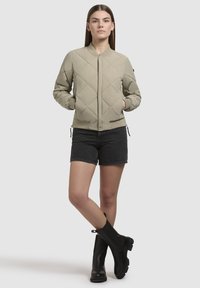 Bomberjack in lichtbeige met diamantgequilte patroon, ribgebreide manchetten en zoom, met zijzakken en een ritssluiting, gecombineerd met zwarte shorts en laarzen.
