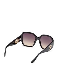 Lunettes de soleil carrées surdimensionnées noires avec verres marron dégradés et logos circulaires dorés sur les branches épaisses, présentées sous un léger angle.