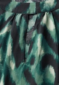 Yksityiskohta laskostetusta kankaasta vihreässä ja mustassa abstraktissa kuviossa, jossa on pieni avainreikä, valmistettu pehmeästä, teksturoituneesta materiaalista.