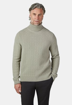 Hombre de mediana edad con pelo gris que lleva un jersey de cuello alto texturizado gris claro y pantalones negros, de pie con una mano en el bolsillo.