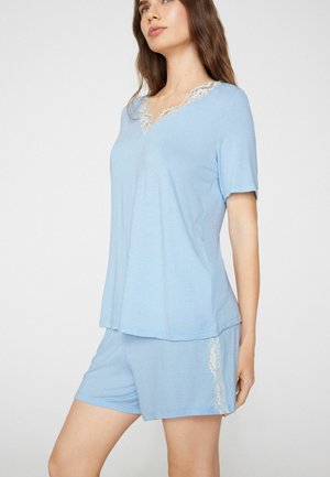 Femme portant un haut bleu clair à manches courtes et un short assorti avec un bord en dentelle blanche autour de l'encolure et sur le côté du short.