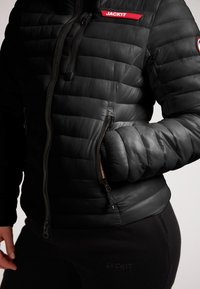 Giacca a vento nera con texture a sandwich orizzontale, chiusura con zip e dettaglio del logo rosso. Caratterizzata da una vestibilità aderente e una comoda tasca.