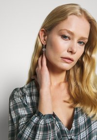Zilveren oorringen met een ronde hanger met ingewikkelde details, gedragen door een model met blond haar, gekleed in een geruite blouse.