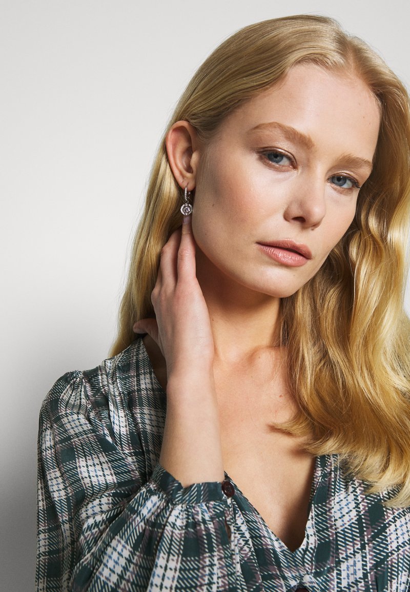 Zilveren oorringen met een ronde hanger met ingewikkelde details, gedragen door een model met blond haar, gekleed in een geruite blouse.