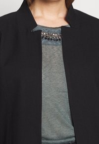 Chaqueta negra con cuello alto, superpuesta sobre una blusa azul claro con un tejido texturizado y tachuelas decorativas a lo largo del escote.