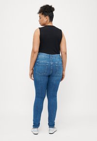 Calças de ganga de cintura alta em azul com uma textura suave, apresentando dois bolsos traseiros e uma silhueta ajustada. Usadas com uma blusa preta sem mangas.