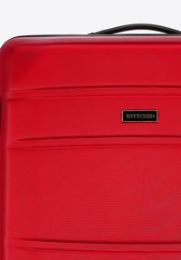 Maleta roja de concha dura con superficie texturizada, acentos negros y un logo de la marca. Presenta líneas horizontales como detalle de diseño y un asa robusta.
