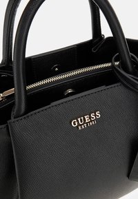 Sac à main synthétique noir avec une finition texturée, logo doré et fermeture zip. Dispose de double poignées et d'un design structuré.