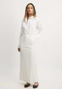 Witte blouse met knopen gecombineerd met een lange wit gestreepte rok. Het model draagt zwarte ballerina's. Gladde stof, aangesloten taille en verticale strepen.