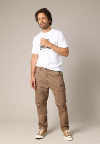 Witte katoenen t-shirt met grafische print, gecombineerd met tan cargo broek met zakken en een ontspannen pasvorm. Sneakers maken de outfit af.