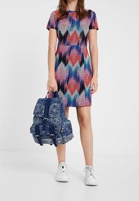 Abito fitted multicolore con motivo a zigzag; zaino in denim con dettagli borchiati, due spallacci e una tasca con zip. Sneakers bianche.