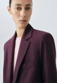Blazer ajusté bordeaux avec revers, poche poitrine simple et texture de tissu lisse. Porté par-dessus un haut rose clair à col rond.
