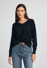 Czarny sweter w serek z długimi rękawami, wykonany z mi ękkiego materiału, z małym czerwonym logo na piersi i luźnym krojem. Noszony z niebieskimi dżinsami.