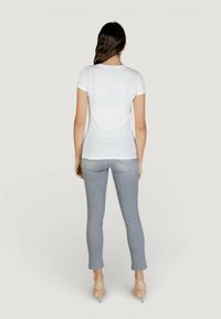 Femme aux longs cheveux foncés portant un T-shirt blanc ajusté, un jean skinny gris clair, et des talons hauts couleur nude, debout de dos sur un fond uni.