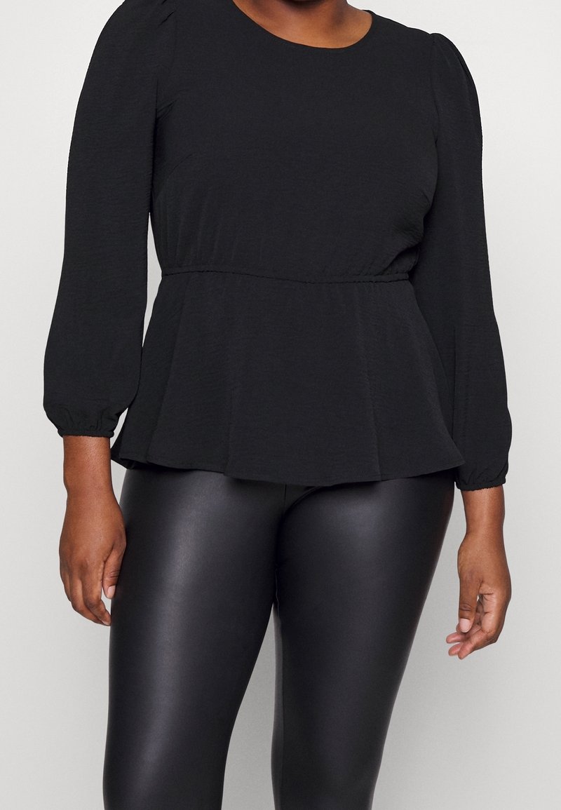 Femme portant un top péplum noir à manches longues associé à un legging noir en simili cuir, debout devant un fond uni.