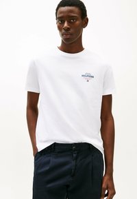 White cotton t-shirt featuring a small blue and red "Hilfiger" logo on the left chest, paired with dark navy trousers. Smooth texture.