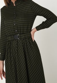 Robe à motif pied-de-poule en vert et noir, avec des manches longues, une fermeture à boutons à l'avant et une ceinture en tissu avec une boucle noire.