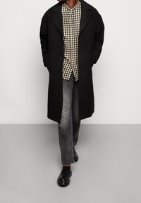 Schwarzer Wollmantel mit Reverskragen, geknöpfter Front, getragen über einem beige- und schwarz karierten Hemd, kombiniert mit grauen Jeans und schwarzen Schuhen.