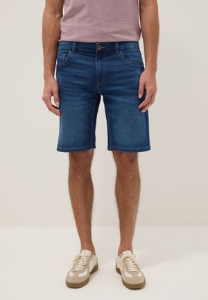 Hombre con pantalones cortos de mezclilla azules, camiseta rosa y zapatillas beige, de pie contra un fondo claro y liso.