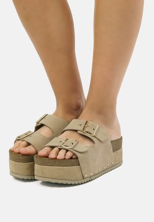 Weibliche Füße, die beige Plateausandalen mit zwei verstellbaren Schnallenriemen und einer dicken Korksohle tragen.